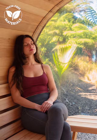A woman relaxes with eyes closed on a wooden bench in a curved wooden room, with sunlight streaming through a large window and a "Toxin-Free Materials" logo in the corner.
