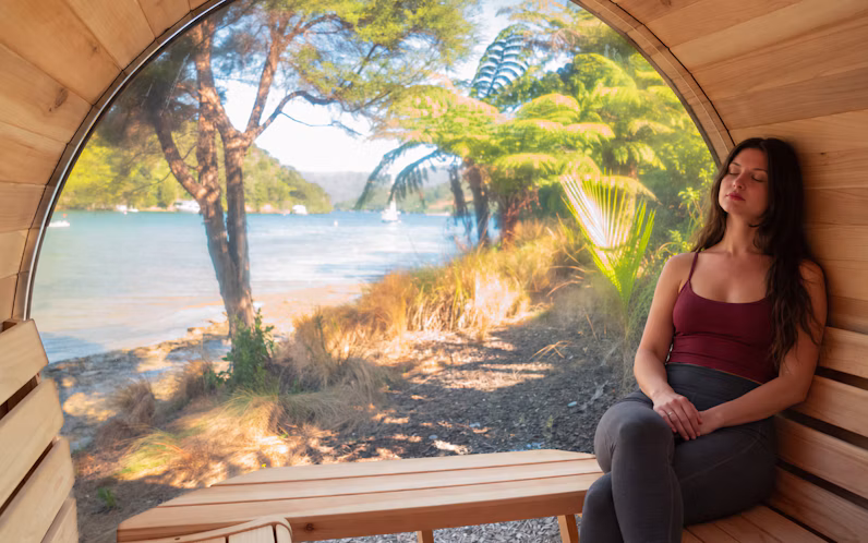 A woman relaxes on a wooden bench inside a barrel-shaped room with a large window overlooking a scenic lakeside landscape.