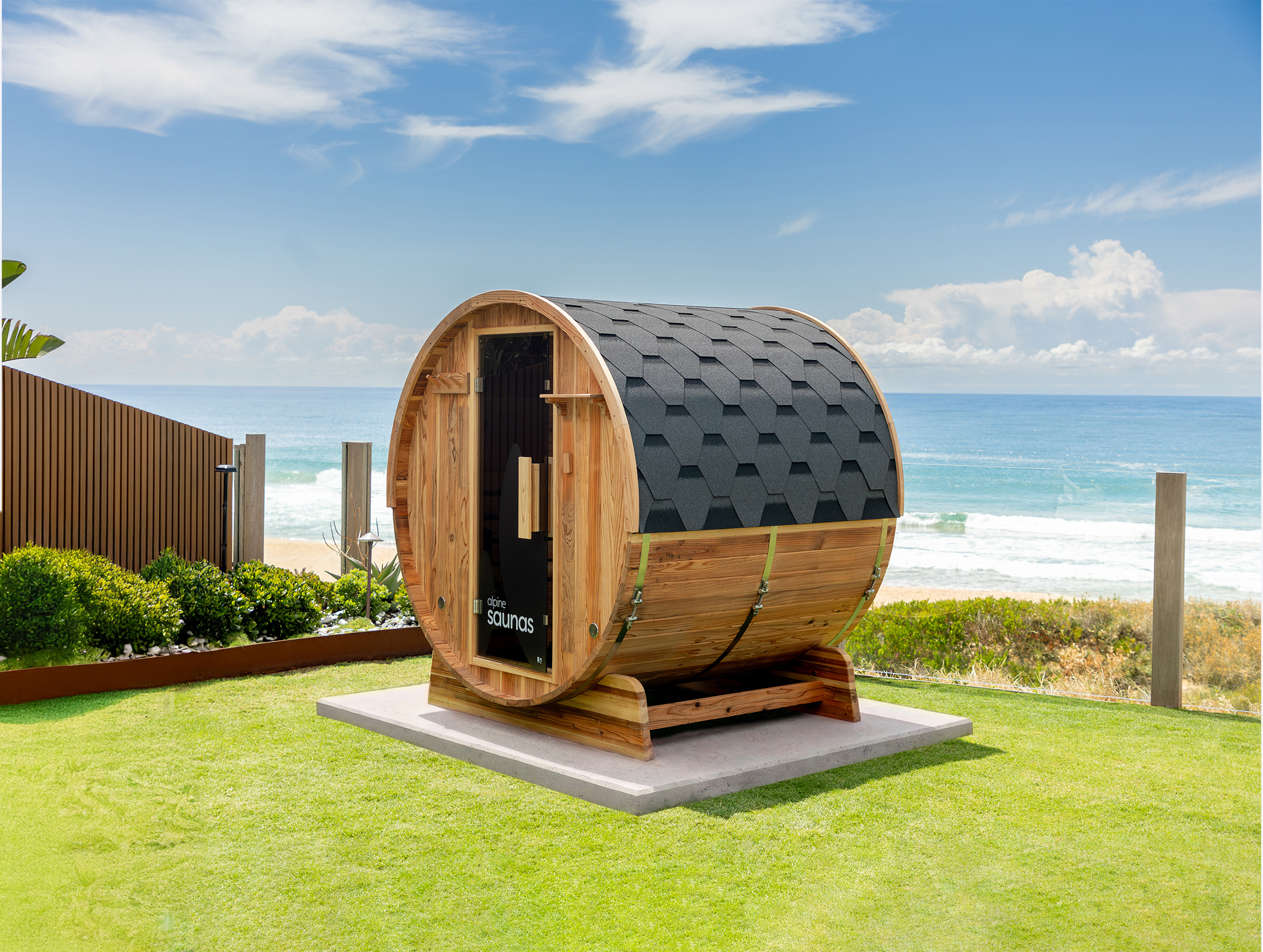 A barrel-shaped wooden sauna with a shingled roof stands on a lawn overlooking the ocean under a blue sky.