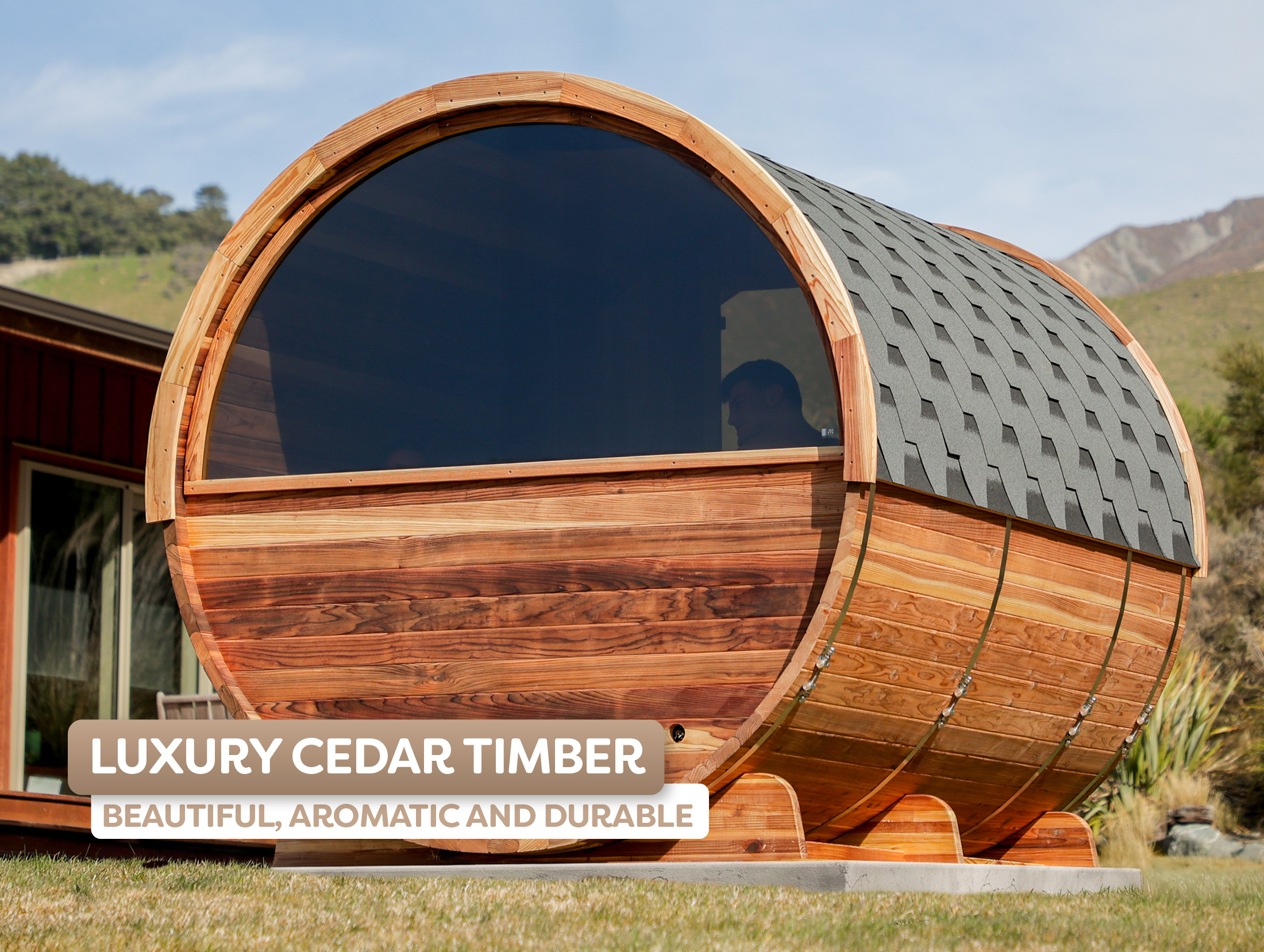 A large barrel-shaped cedar timber structure with a shingled roof sits outdoors beside a house, with a person visible inside.