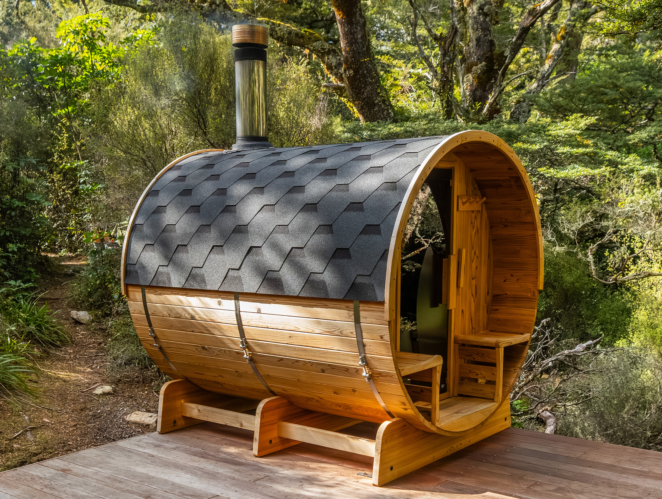 A wooden barrel-shaped structure with a shingled roof and chimney stands on a wooden deck in a lush forest setting.