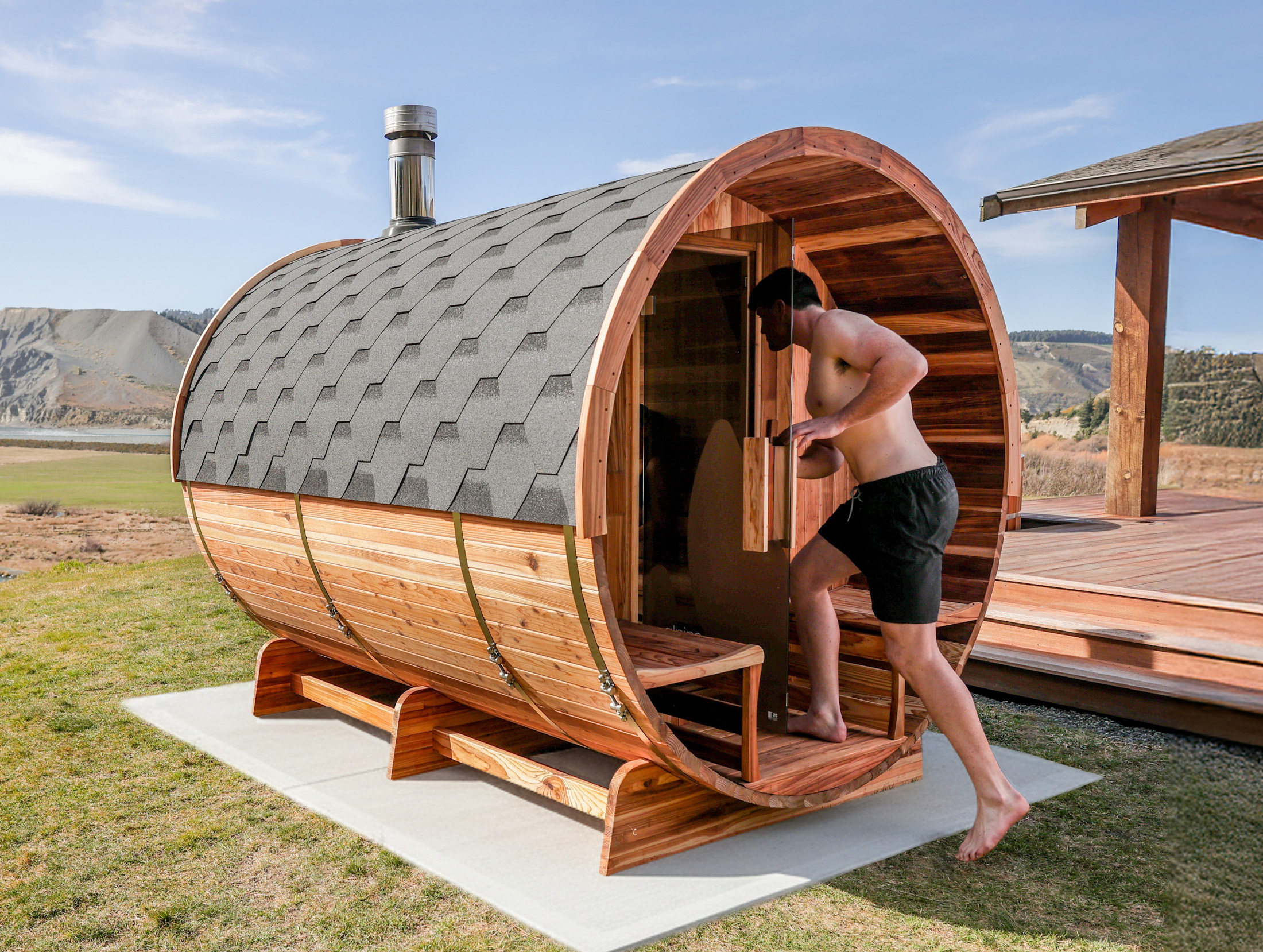 A man in swim shorts is entering a barrel-shaped wooden outdoor sauna situated on a grassy area with mountains in the background.