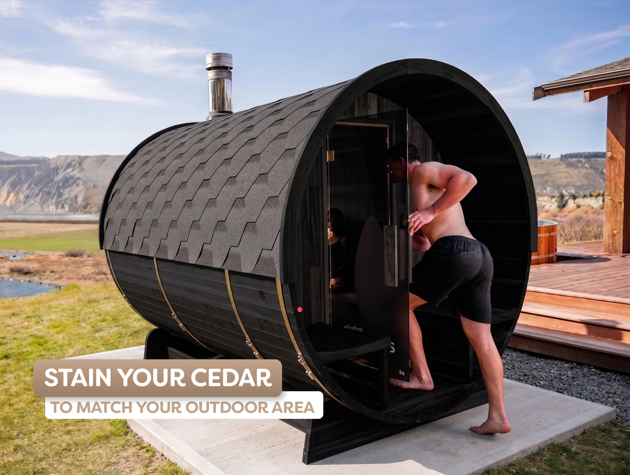 A shirtless man in shorts enters a barrel-shaped outdoor sauna with dark stained cedar wood, set in a scenic landscape.