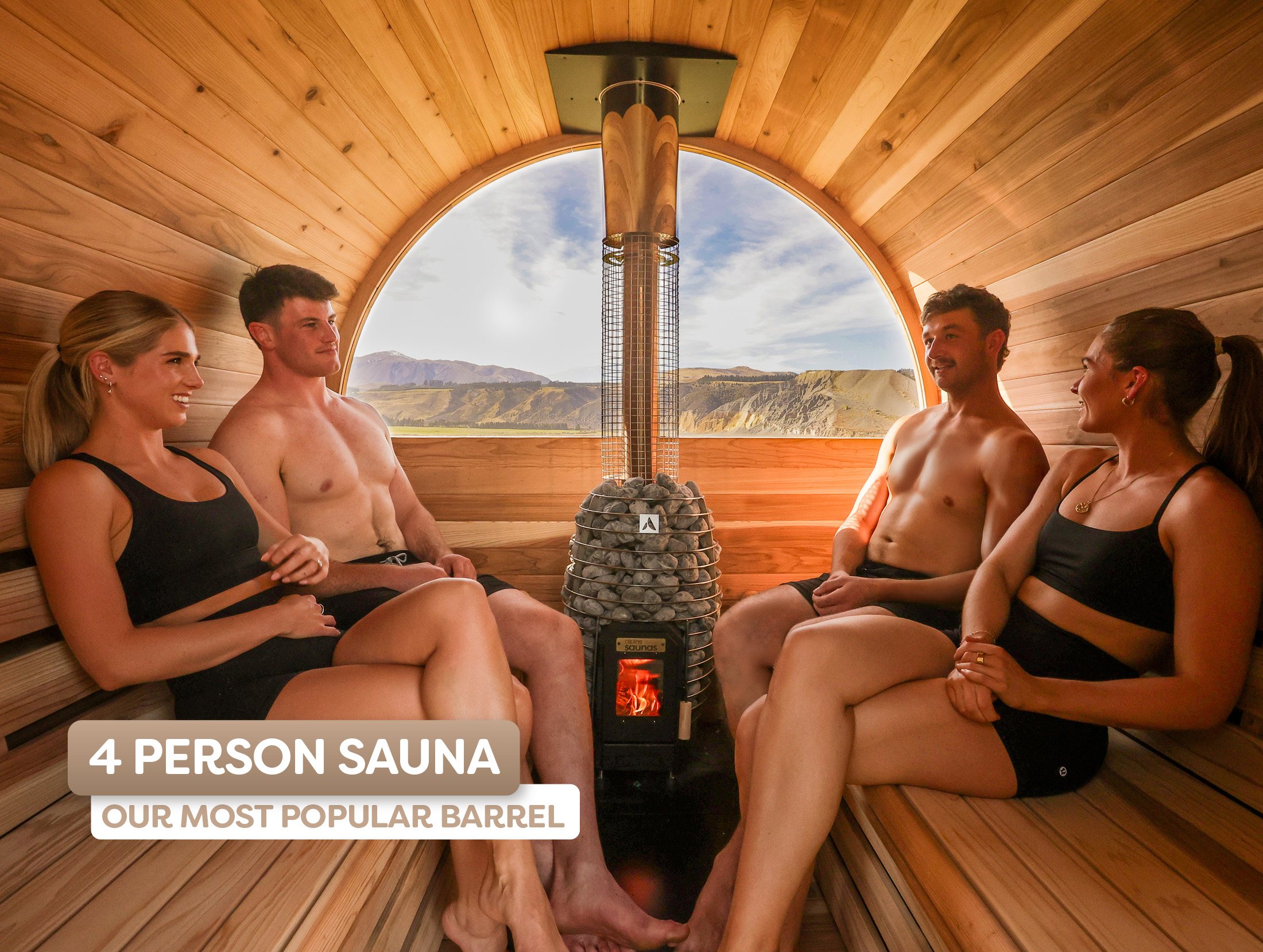Four people in swimwear sit inside a wooden barrel sauna with a central heater, enjoying the warmth and mountain views through a large window.