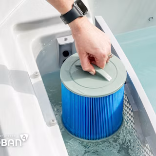 A hand wearing a smartwatch lifts a blue cylindrical filter cartridge from the water in a hot tub; the word "Microban" is visible on the tub's edge.