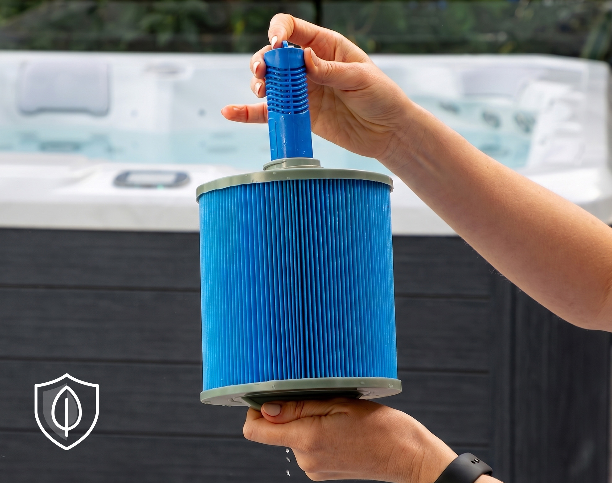 A person holds a blue cylindrical hot tub filter cartridge with water dripping from it, with a hot tub in the background and a leaf shield icon in the corner.