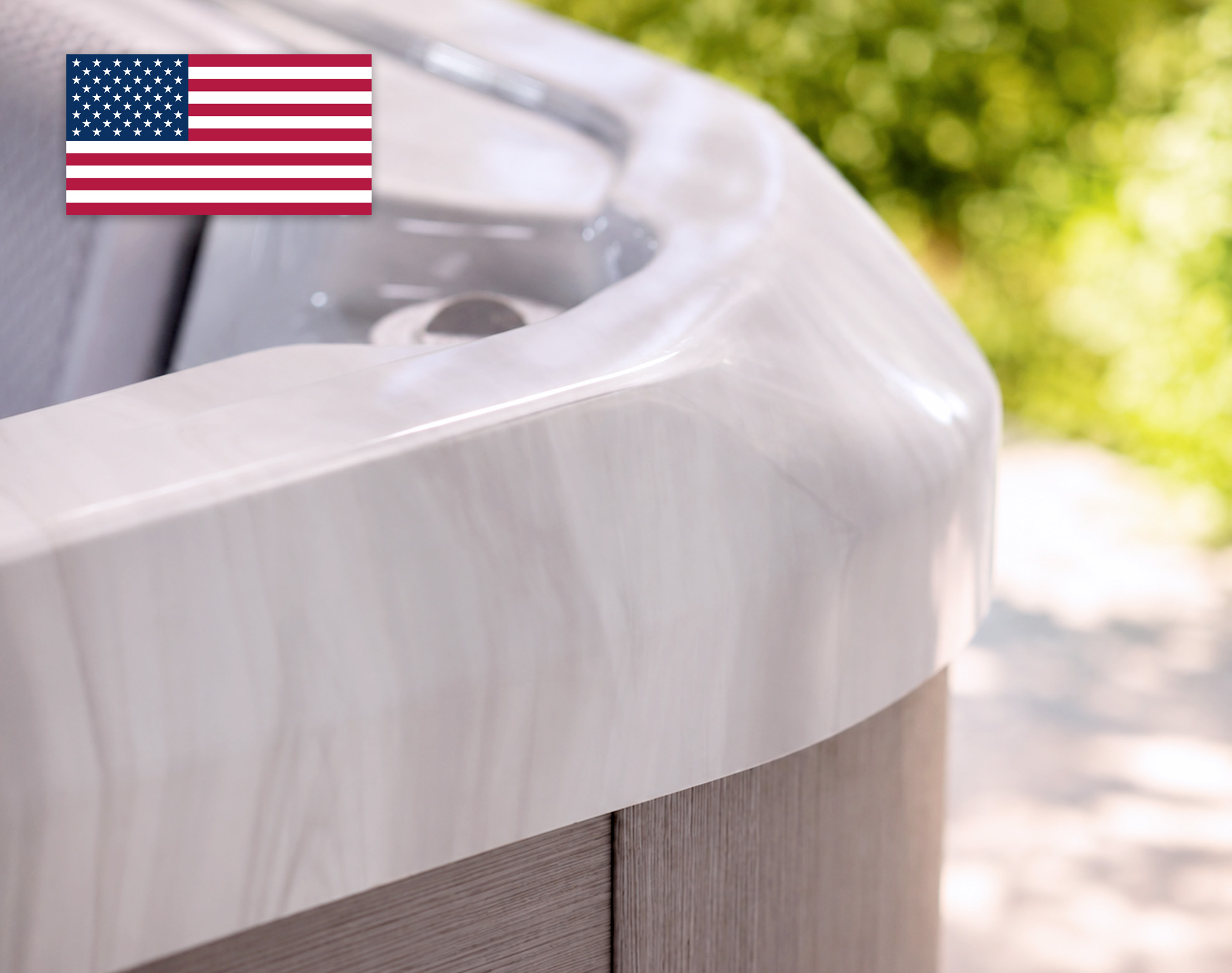 Close-up of a platinum acrylic hot tub shell with a blurred background of greenery and an American flag in the upper left corner.