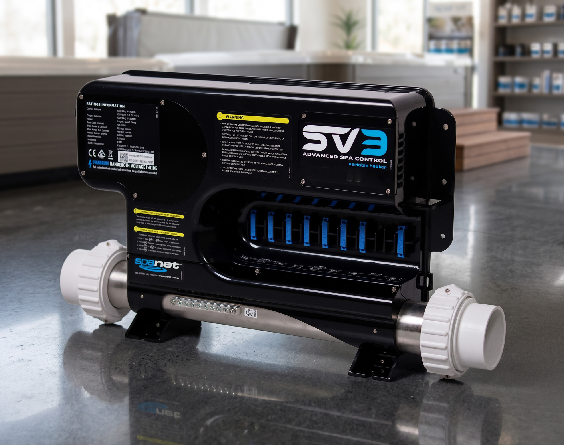 A black and silver spa heater unit labeled "SV3 Advanced Spa Control variable heater" sits on a reflective floor, featuring control labels, warning stickers, and white pipe connectors at each end.