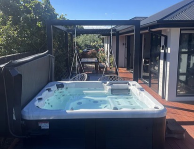 A square outdoor spa pool filled with water is positioned on a wooden deck beside a modern house, with a pergola and garden in the background.