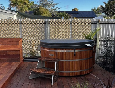 A round wooden outdoor tub with a cover and small steps, set on a timber deck in a fenced garden.