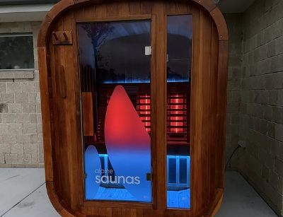 A small wooden sauna with a glass door and interior illuminated by red and blue lights is positioned outdoors against a brick wall.