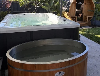A round wooden tub filled with water is in the foreground, with a larger rectangular tub and a wooden outdoor structure in a garden setting.