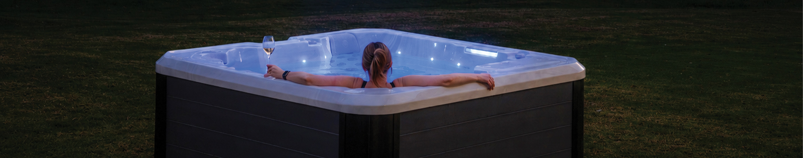 A person relaxes in an illuminated outdoor spa pool at dusk, holding a glass of wine.